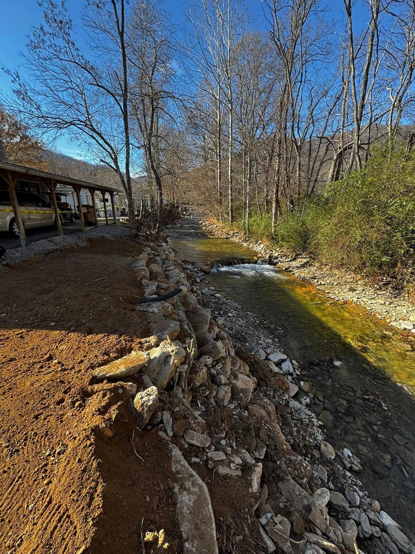 Restored creek bank with a stone-armored toe, reshaped soil bank and a new drain line, running alongside a gravel parking area under bare winter trees.