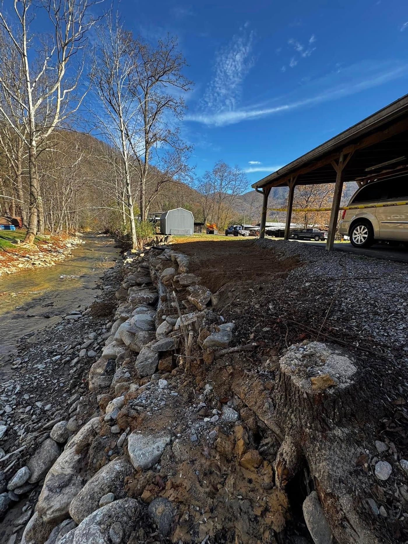 Storm-scoured creek bank with an undercut edge, exposed tree stump and loose rock, threatening the gravel parking area and carport above it.