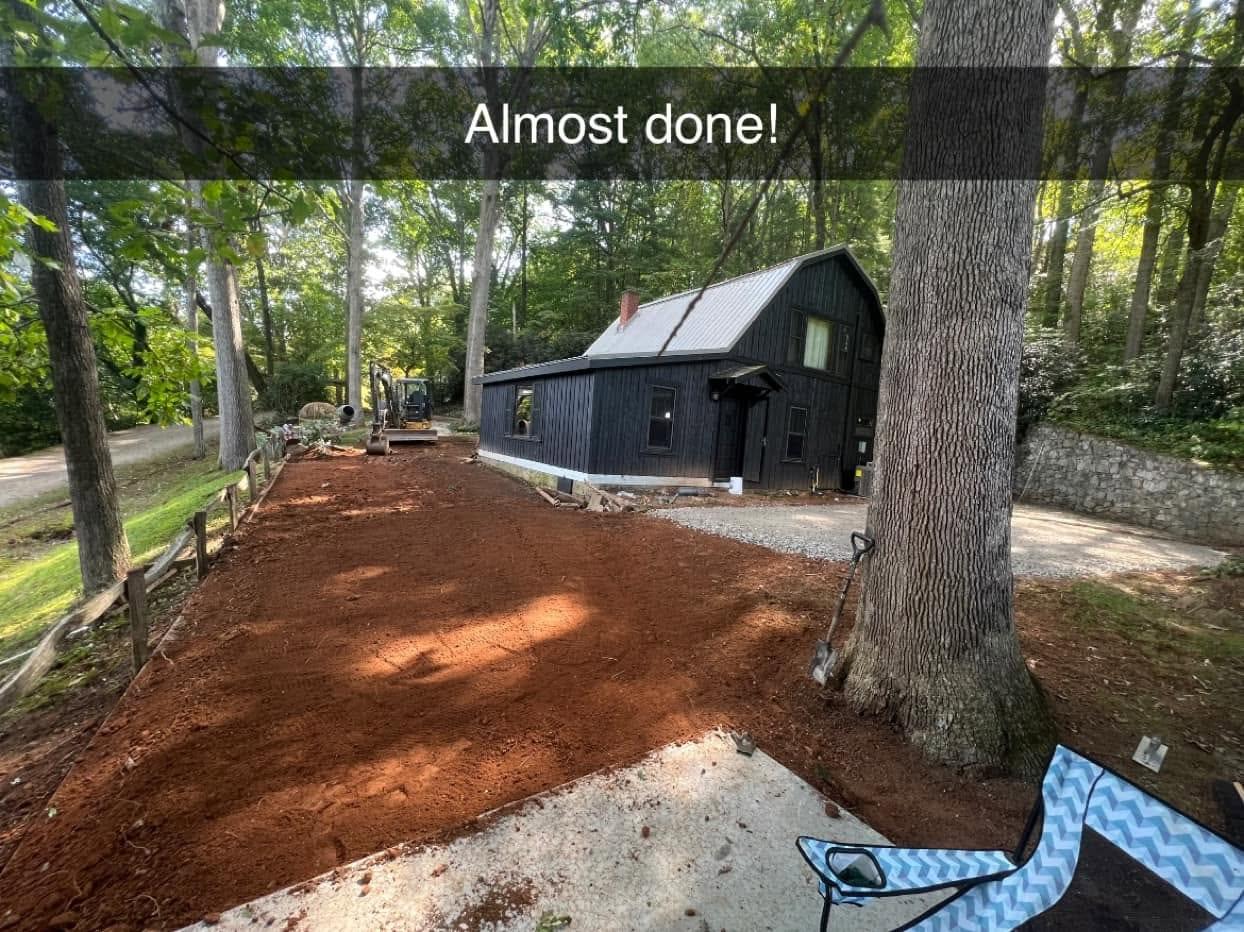 Freshly graded red-clay driveway and building pad beside a black barn-style cabin in the woods, with a compact excavator parked at the far end of the cut.