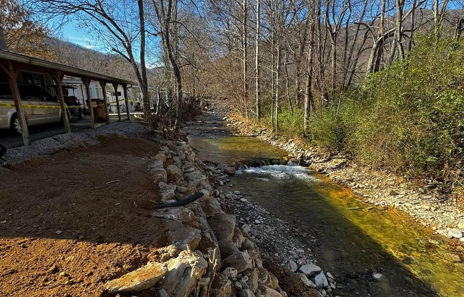 A restored mountain creek running past a stone-armored bank and freshly graded ground, with a blue-sky ridgeline rising behind the bare winter trees in western North Carolina.