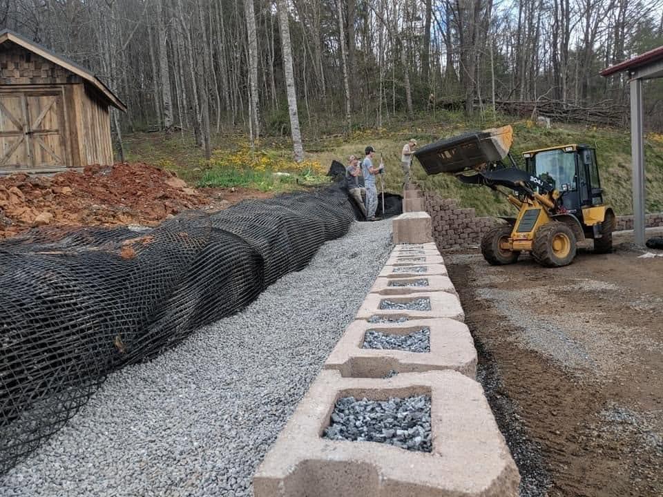 LWM Consulting crew installing a geogrid-reinforced segmental block retaining wall with washed drainage stone, while a wheel loader places fill behind them.